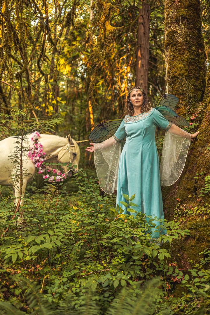Woman dressed as a fairy standing beside her unicorn-adorned horse in a whimsical forest portrait session.
